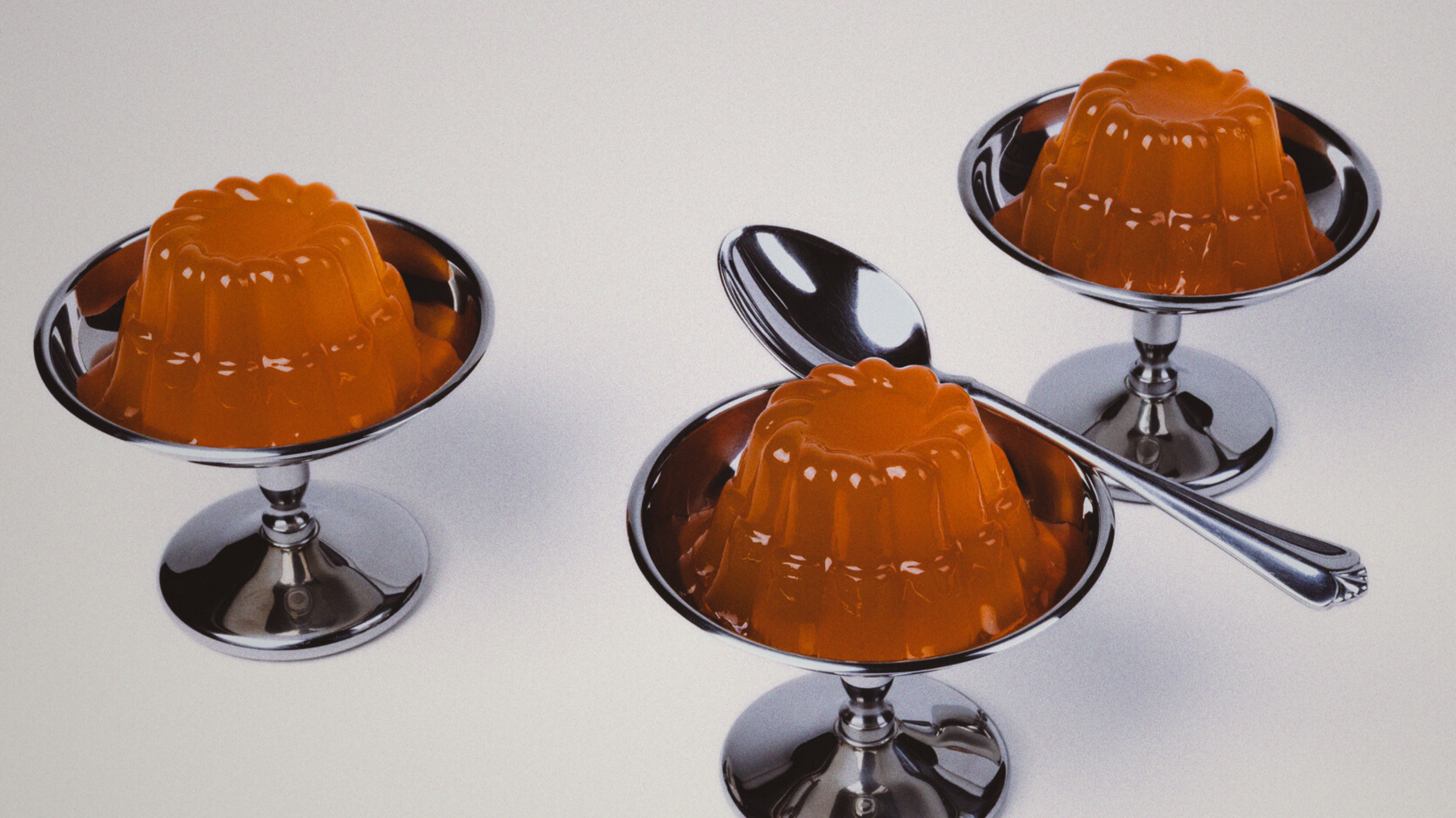 Three orange gelatin molds on metallic stands with spoons on a white background