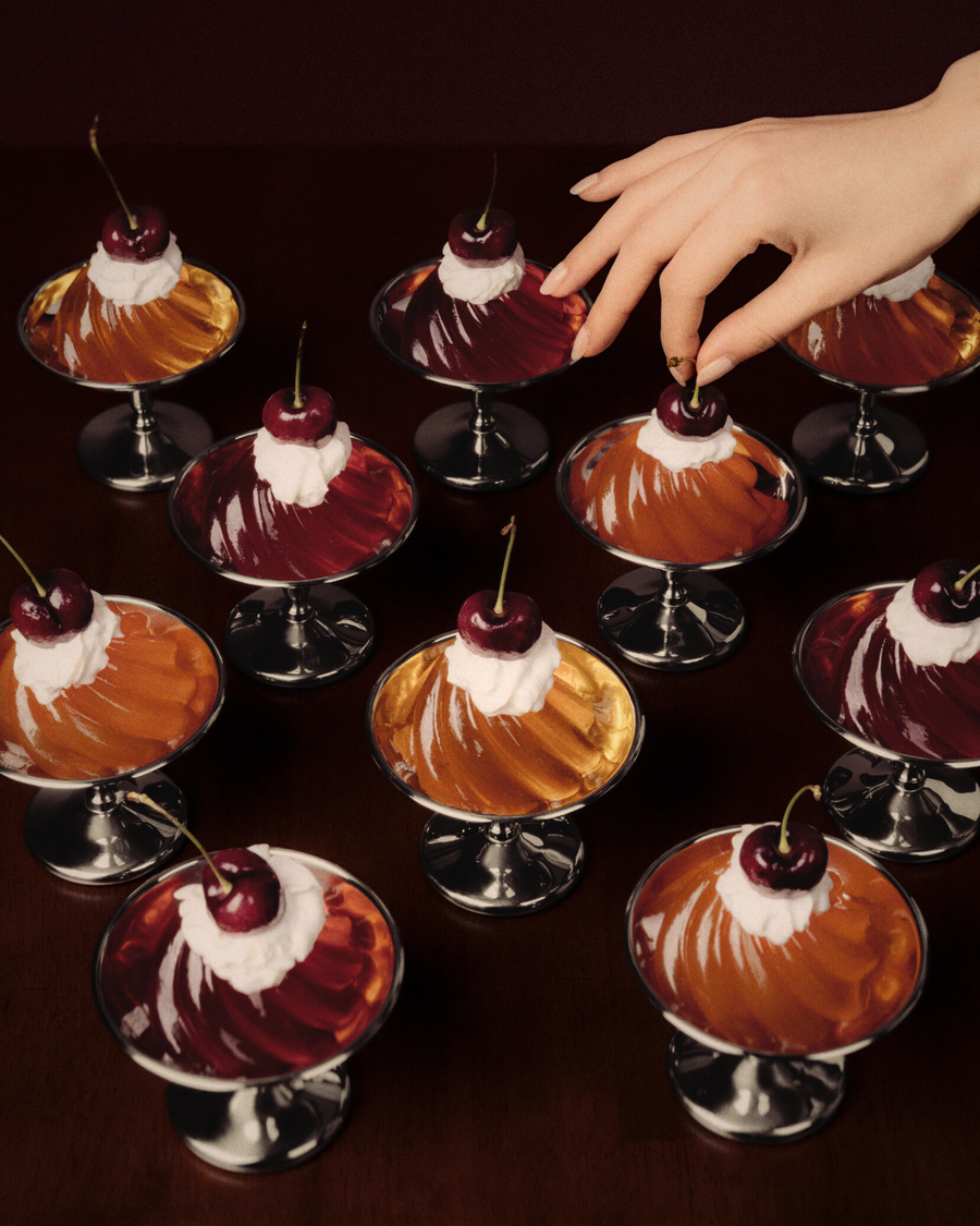 Dessert glasses with cherry-topped multi colored gelatin on a dark surface, with a hand reaching in.